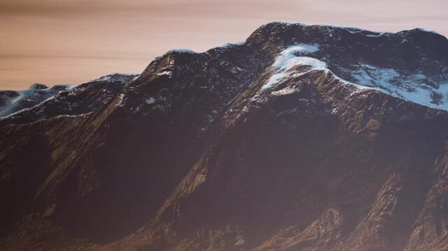 Snow covers the high peaks of mountains as the sun sets in the background. The fading light casts a warm glow on the rocky surface and snow.