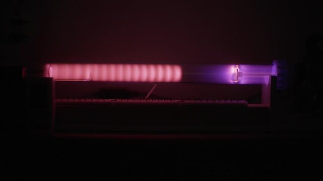 Close up view of a pulsed plasma test displaying glowing pink and purple striations inside a glass vacuum tube in a dark laboratory setting.