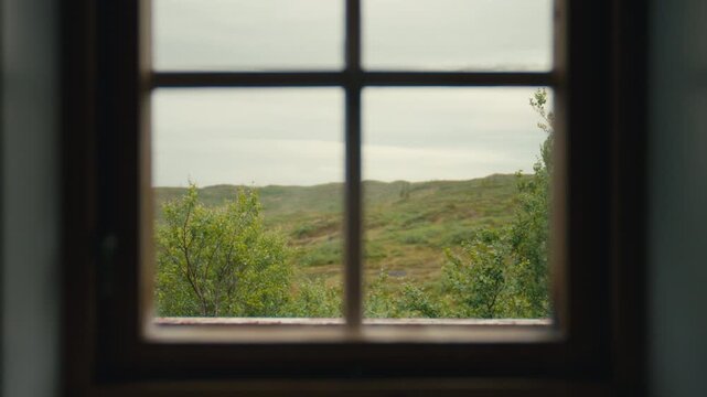 A soft mountain landscape is seen through a rustic window frame, where muted daylight, distant hills, and the sheltered indoor viewpoint create a quiet scene of stillness, distance, and rural calm