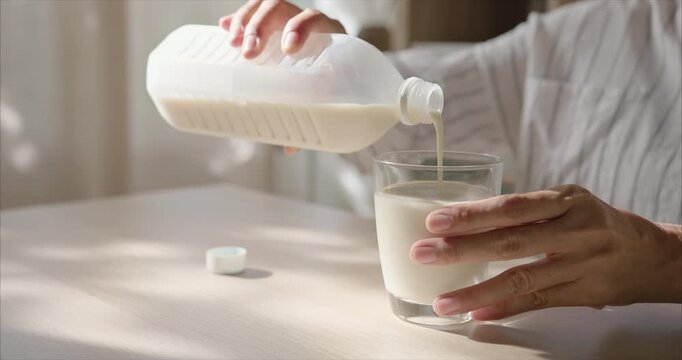 Close up of Asian Woman Hands Pouring Soy Milk into Glass on Table in Cozy Living Room with Warm Morning Sunlight, Healthy Breakfast Routine