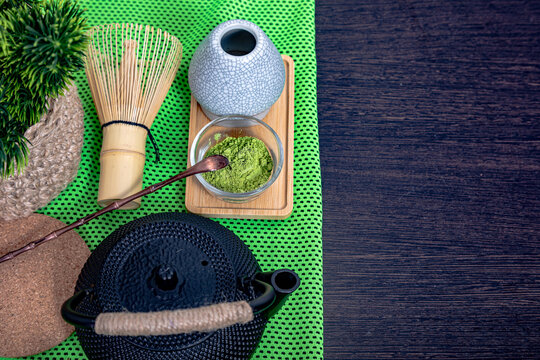 Matcha powder with bamboo whisk and black iron teapot on textured green mat. Traditional Japanese tea ceremony equipment. Healthy organic green tea preparation concept, top view