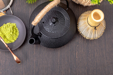 Fototapeta premium Matcha tea powder with bamboo whisk and black iron teapot on dark wooden table. Japanese tea ceremony preparation with organic green tea leaves. Healthy natural lifestyle tradition