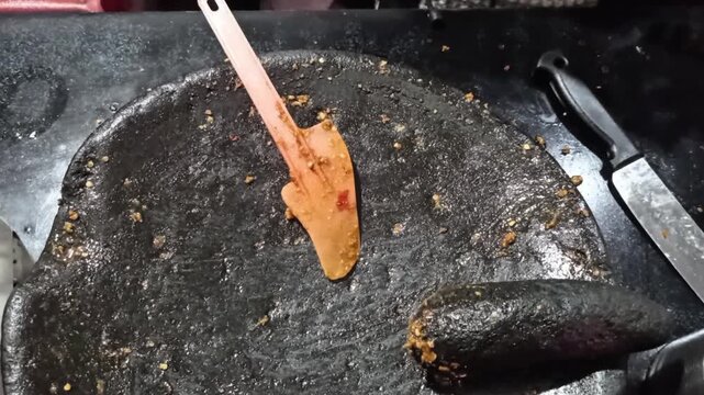 Close-up of hands using a spatula to scrape traditional Indonesian sambal chili paste from a large black stone mortar into small plastic containers.