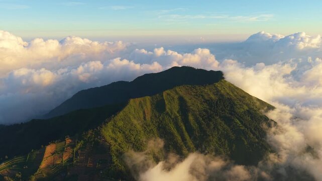 Aerial view of Mount Sindoro and Sumbing with terraced fields and lush green slopes surrounded by clouds at golden hour in Kecamatan Kedu, Jawa Tengah, Indonesia.