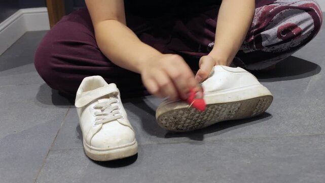 Hands scrub dirt from a white sneaker while the person sits on a gray tile floor.