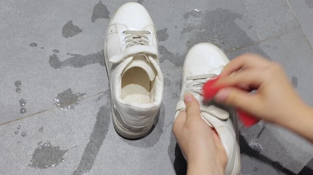 Hands wipe a white sneaker with a red sponge in a close indoor shoe care view.
