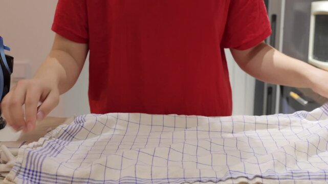 A woman unfolds and spreads a checked kitchen towel on an ironing board before pressing it with a steam iron.
