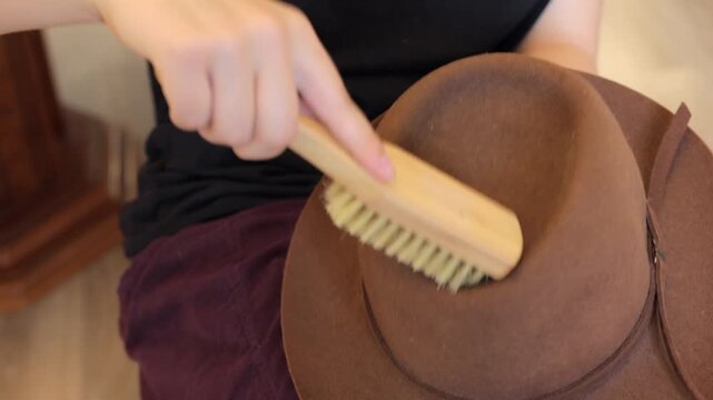 Close-up hands brush a brown felt hat indoors, focusing on dust removal, texture, and careful wardrobe maintenance.