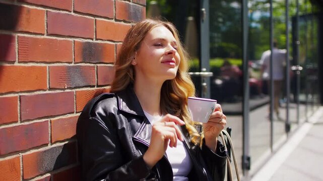 Fashionable woman sitting by an outdoor cafe table by a brick wall and drinking a cup of hot coffee