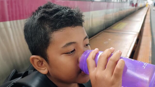Close-up of an Asian boy drinking from a purple water bottle on a train station platform, staying hydrated next to a commuter train during his travel journey.