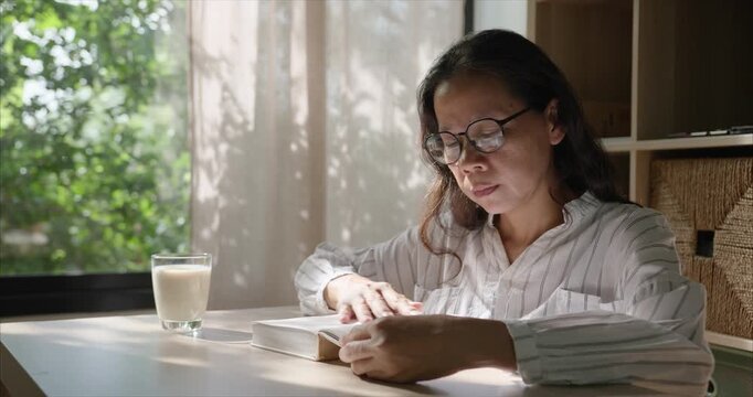 Middle Aged Asian Woman with Glasses Reading Book in Living Room, Cozy Home Interior.	