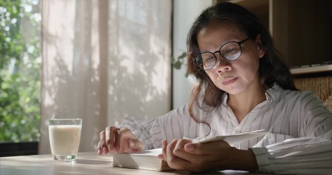 Middle Aged Asian Woman with Glasses Reading Book in Living Room, Cozy Home Interior.