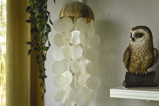 Cozy home interior decoration with a capiz shell wind chime hanging near a decorative owl figurine on a shelf. Boho style apartment details, soft indoor lighting.