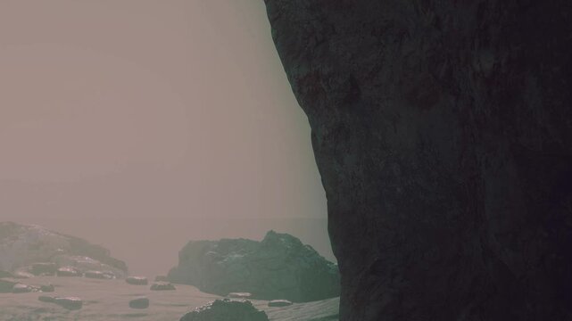 A person walks on a beach covered with rocks while fog surrounds the area. A large cliff stands on one side of the beach, adding to the scene.