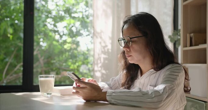 Side view. Asian Woman with Glasses Using Smartphone in Living Room, by Window with Warm Morning Sunlight.