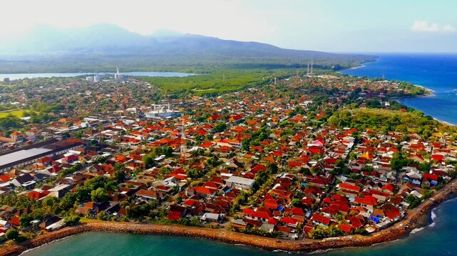 Aerial view of the coastal town of Gilimanuk Bay with red-roofed houses and lush greenery along the shoreline in Kecamatan Melaya, Bali, Indonesia.