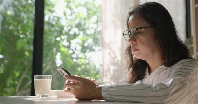 Side view. Asian Woman with Glasses Using Smartphone in Living Room, by Window with Warm Morning Sunlight