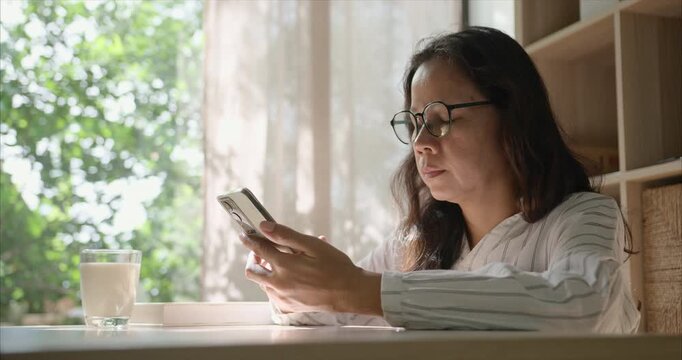 Side view. Middle Aged Asian Woman with Glasses Using Smartphone in Living Room,  by Window with Warm Morning Sunlight, Home Interior Backdrop