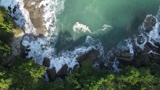 Aerial view of crashing waves against rocky cliffs and lush green vegetation along the coastline in Kebumen, Central Java, Indonesia.