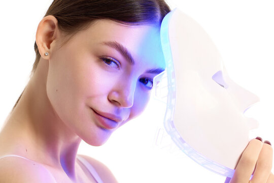 Woman smiling beside blue LED face mask for beauty treatment. Concept of selfcare, antiaging skincare, acne therapy, beauty gadget ads and home spa routine.