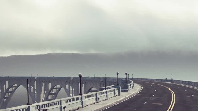A road winds towards a large bridge. The sky is mostly cloudy with fog covering the hills. Street lamps line the road as it curves gently. The scene appears quiet and empty.