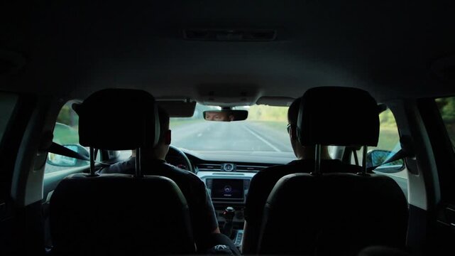 Two men driving car on highway, view from backseat showing driver and passenger, travel concept, road trip journey, vehicle interior perspective during daytime, journey on open road