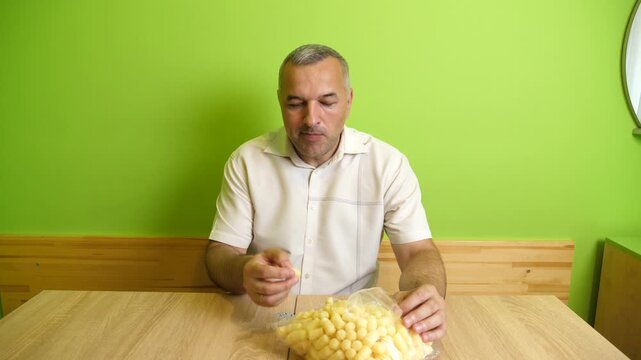 A man sits at a table eating corn puffs, taking them from a transparent bag indoors against a green wall.