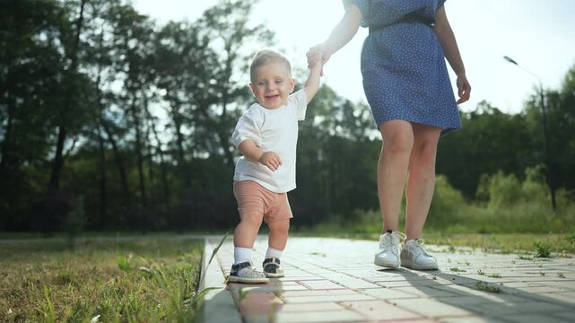 Mother and happy toddler in park. Toddler holding mother hand walking in park. Happy family moments outdoors. Toddler learning to walk in park with mother. toddler and mother bond in park. first steps
