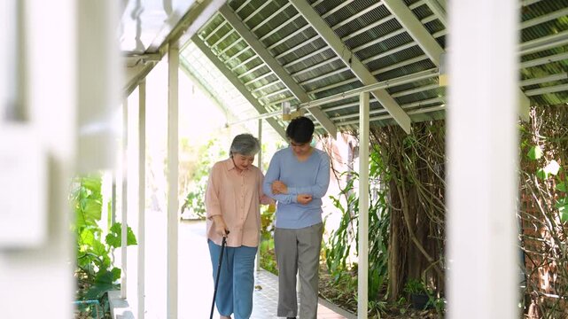 Adult son accompanies his elderly mother using walking cane as they walk toward restaurant together. Family care, support, healthy aging, independence and multigenerational lifestyle concept.