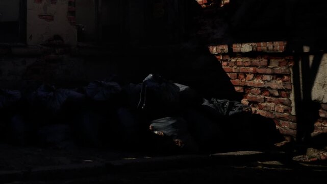 People throw away objects in a dark space next to a crumbling brick wall. Bags are piled up and the light casts shadows on the ground. This spot shows signs of neglect and urban decay.