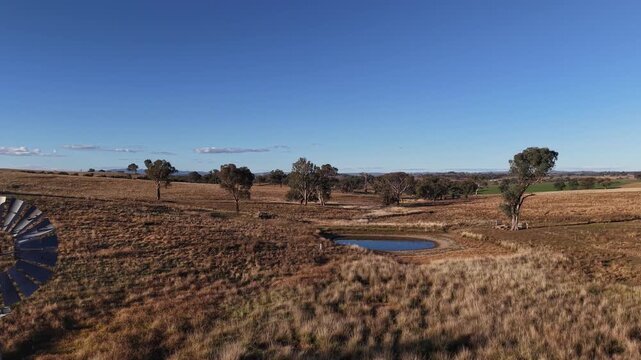 Aerial view of a traditional metal windmill standing in a dry grassy field next to a small pond with scattered eucalyptus trees in Canowindra, New South Wales, Australia.