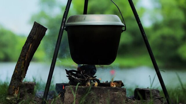 Metal pot suspended over campfire by tripod cookout outdoors. Cooking activity with smoke, fire, and glowing embers against blurred green background of river and trees.