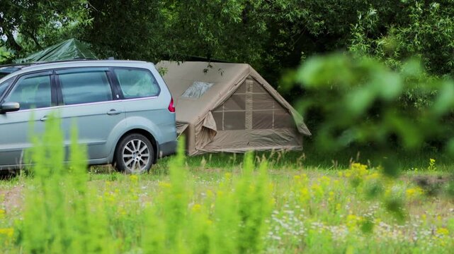 Light blue minivan vehicle parked near large tan canvas camping tent setup in lush green field, abundant trees and wildflowers surround outdoor summer recreation area, copy space