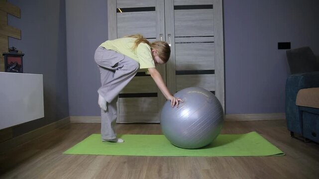 Young caucasian girl performing back stretch exercise with stability ball on green yoga mat indoors, practicing fitness routine at home, enhancing flexibility and core strength.