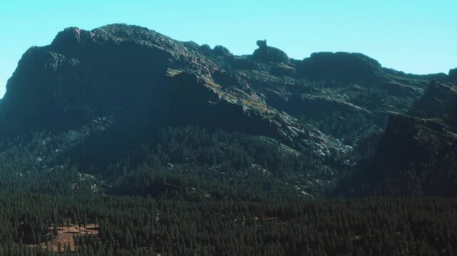 A mountain range rises sharply against a clear blue sky. Green trees cover the lower slopes. The sunlight highlights the rugged surface of the mountains during daytime.