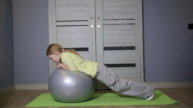 Young girl balancing on large exercise ball performing plank exercise in home room, engaging core muscles for fitness and strength training, developing stability and physical condition.