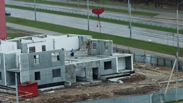 Construction of multistory building using concrete panels by construction workers under large red crane hook overhead, urban development project next to highway road infrastructure.