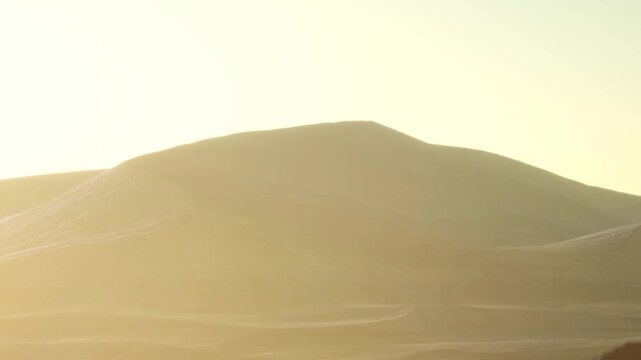 The sun rises over a mountain range creating a warm glow on the sandy hills. The scene shows the vast desert area with soft shadows on the terrain.