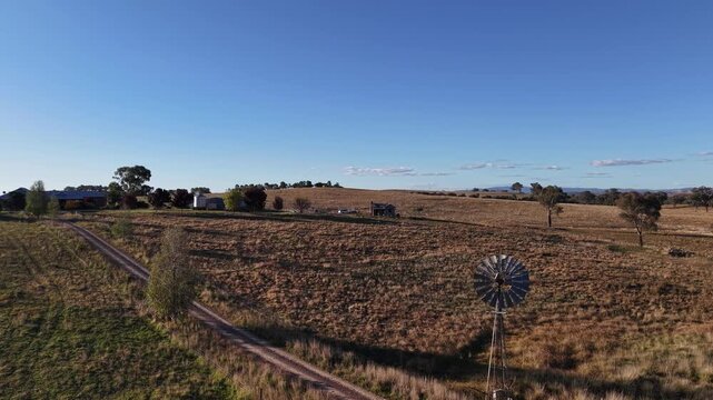 Aerial view of a rural landscape featuring a classic windmill, dry fields, and a dirt road in Canowindra, New South Wales, Australia.