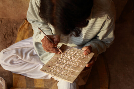 View of a craftsman carving intricate floral patterns into a tile with a small tool, showcasing traditional pottery techniques in Hala, Sindh, Pakistan.