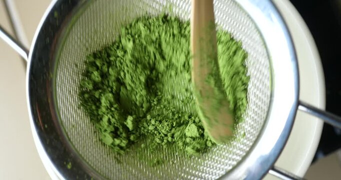 Fine Powder Sifting: A close-up view of vibrant green powder, likely matcha, being sifted through a fine-mesh strainer, showcasing its delicate texture and preparation for culinary use.