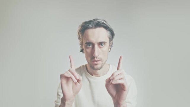 Serious young man in white sweater raising both index fingers, emphasis gesture, isolated on neutral background, communication and presentation concept