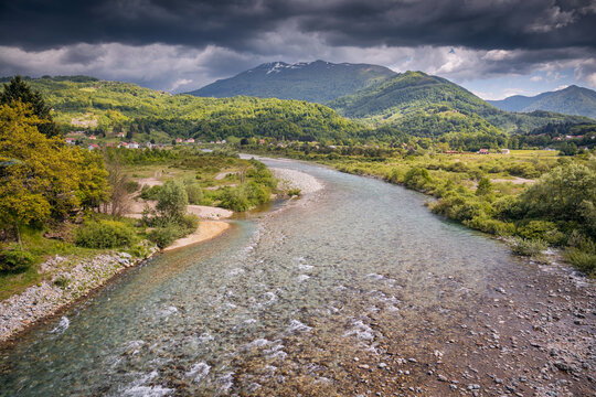 River with clear water running through a lush valley, surrounded by green hills and distant mountains under dark storm clouds