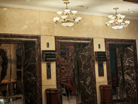 Interior view of a luxury hotel lobby featuring polished marble wall panels, decorative ceiling chandeliers, and modern elevator doors. The design emphasizes elegance, symmetry