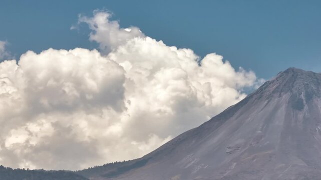 Reveal shot Volcan de Colima (Fire Volcano) with steep volcanic slopes, surrounded by white cumulus clouds and blue sky near Comala, Colima, Mexico