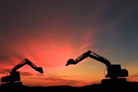 Two Crawler excavators silhouette are digging the soil in the construction site ,with bucket lift up on  sky sunset background.
