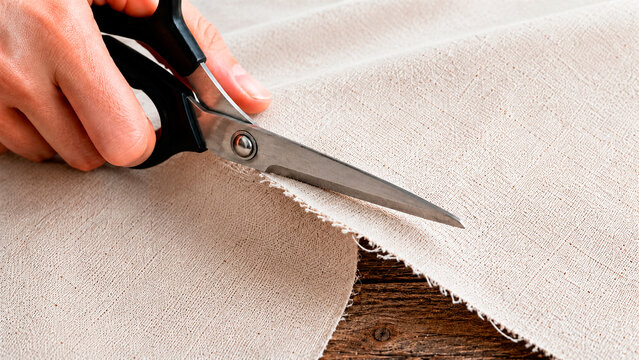 Close up of hands cutting light beige fabric with black scissors. Person tailoring linen cloth on a rustic wooden table. Handmade craft and sewing concept