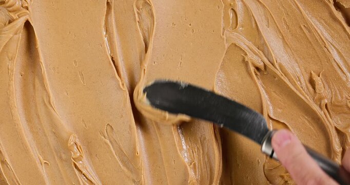 Creamy peanut butter texture background. Top view of a spoon spreading smooth peanut butter across the full frame, creating flowing lines and organic patterns