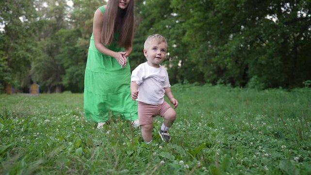 Mother help toddler on grass. Family enjoying outdoor moment. Toddler takes first step on grass. Family bonding during first step. Mother provides help for toddler. Toddler learns to walk with help.