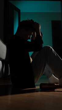 Vertical video silhouette of a man sitting on the floor in a dark room, holding a bottle to his head, depicting struggles with depression, addiction, and loneliness
